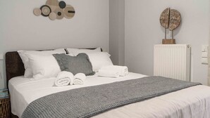 Una cama cuidadosamente tendida con una manta gris, almohadas blancas y un plaid gris. Hay una obra de arte en la pared sobre la cama y un radiador a la derecha.