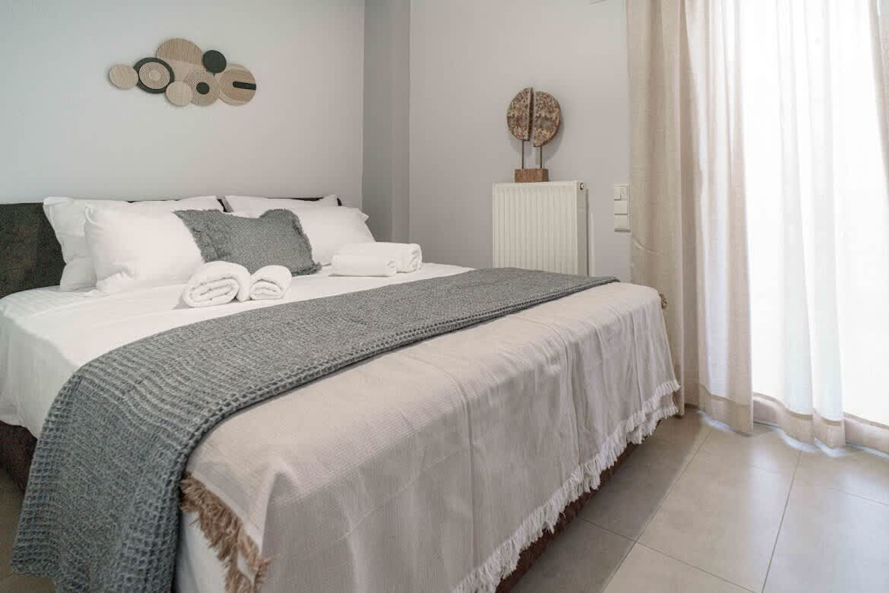 Una cama hecha con cuidado, con una manta gris, almohadas blancas y un edredón blanco. Hay un radiador blanco en la pared y un objeto decorativo redondo encima.