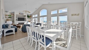 A spacious living area with a dining table, white chairs, and a fireplace. There are large windows offering a view of the ocean.
