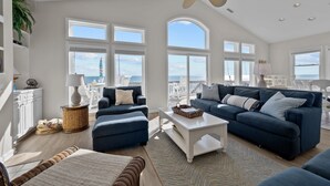 A spacious living room with two blue sofas, a white coffee table, and a large arched window offering a view of the sea.