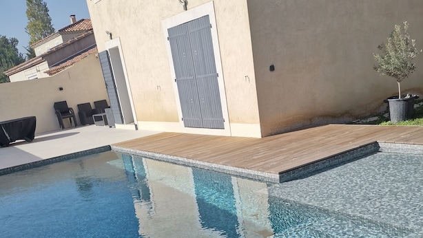 Una piscina con borde de azulejos, cubierta de madera y un edificio con persiana cerrada y mobiliario exterior.