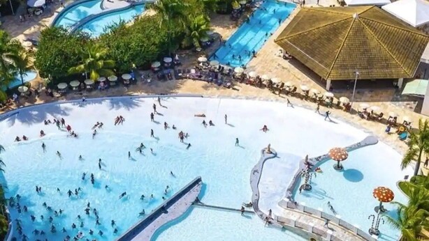 A large outdoor swimming pool area with multiple smaller pools, surrounded by palm trees and shaded by a large thatched structure. - Ap Completo Parq. Aquát. Incrível Grátis (Caldas Novas)