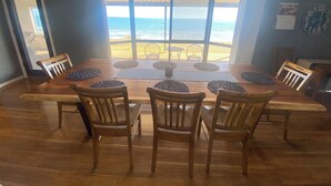 A dining area with a large wooden table and chairs, a view of the ocean through large windows, and a clock on the wall. - Hendys 346 - Absolute beachfront! Excellent sea views! (Point Turton)
