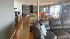 A modern living room with a leather sofa, a dining area with wooden furniture, and a kitchen with white cabinets and a stove. - Hendys 346 - Absolute beachfront! Excellent sea views! (Point Turton)