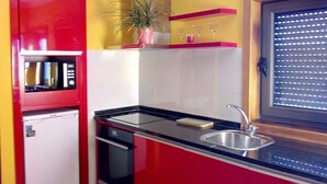 Cocina pequeña con armarios rojos, encimera negra, pileta de acero inoxidable y una ventana con persianas.