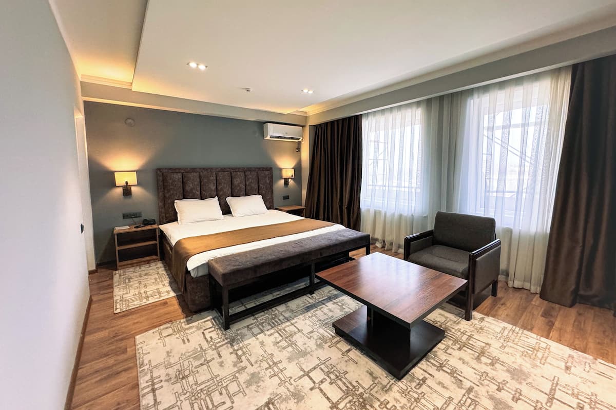 A modern hotel room with a large bed, a bedside table, a chair, a coffee table, and a patterned rug.