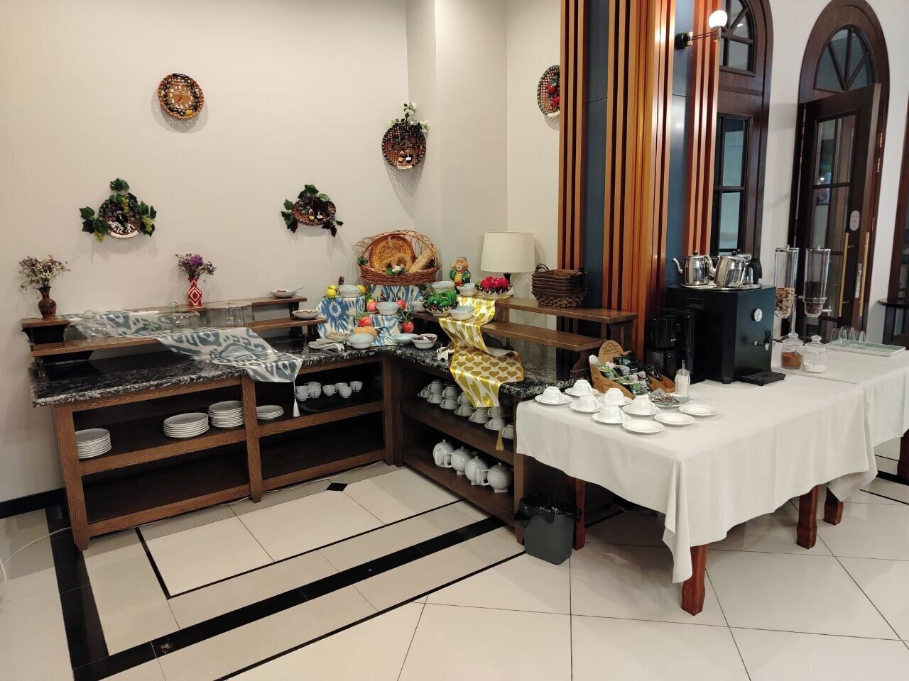 A dining area with a buffet setup, including a variety of dishes and utensils on a table, surrounded by shelves with more dishes and a decorative wall.
