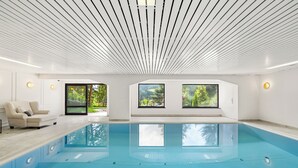 Piscina interior moderna com vista ampla para o exterior através de grandes janelas, área de estar com sofá e teto com padrão linear único.