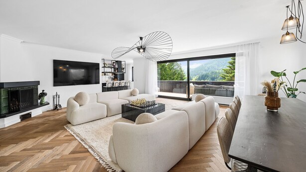 Uma sala de estar moderna com um sofá, uma mesa de centro com peças de xadrez, lareira e uma grande janela com vista para o verde.