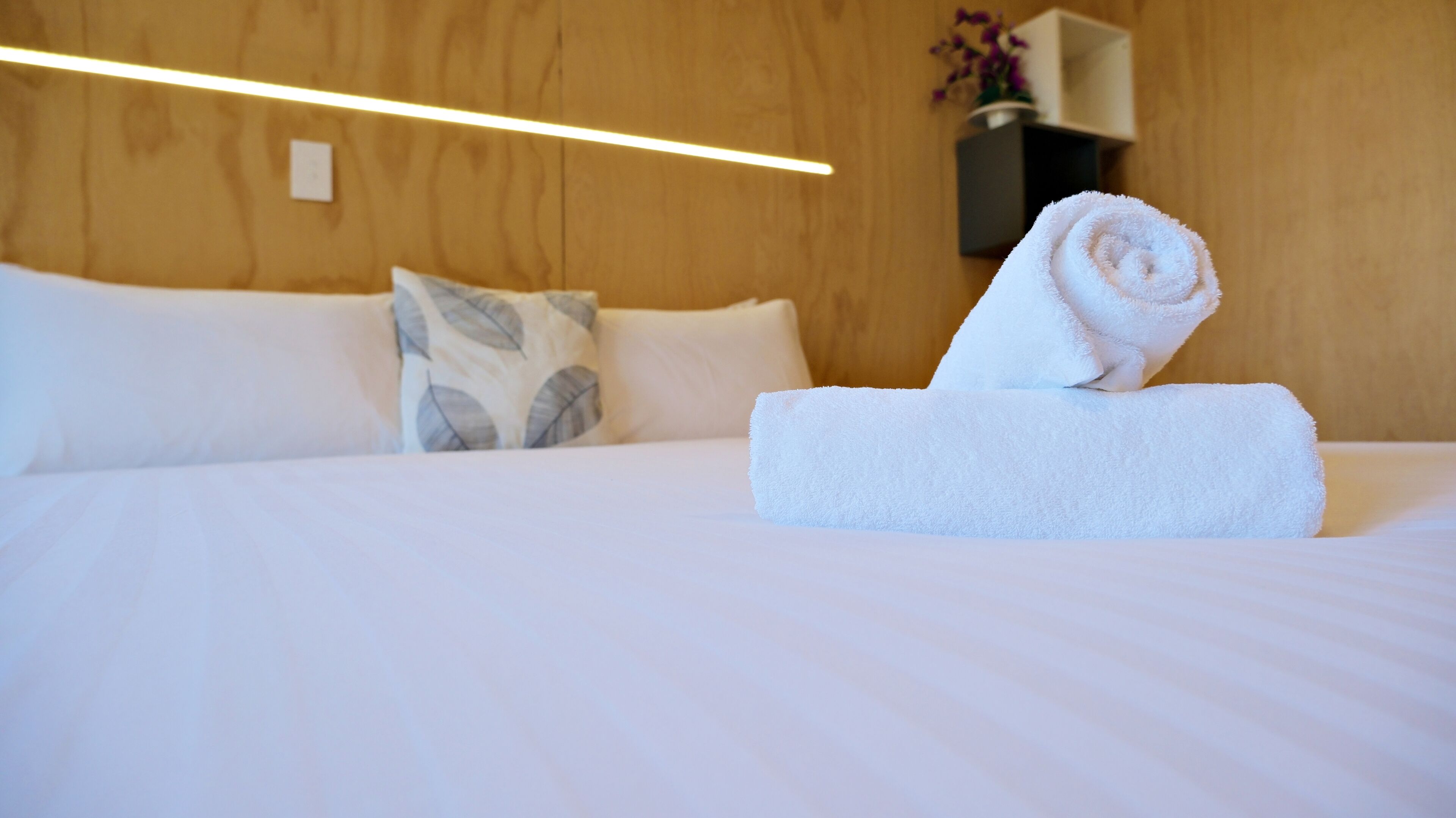 A neatly made bed with a rolled white towel, a pillow with a leaf pattern, and a wall-mounted vase with flowers.