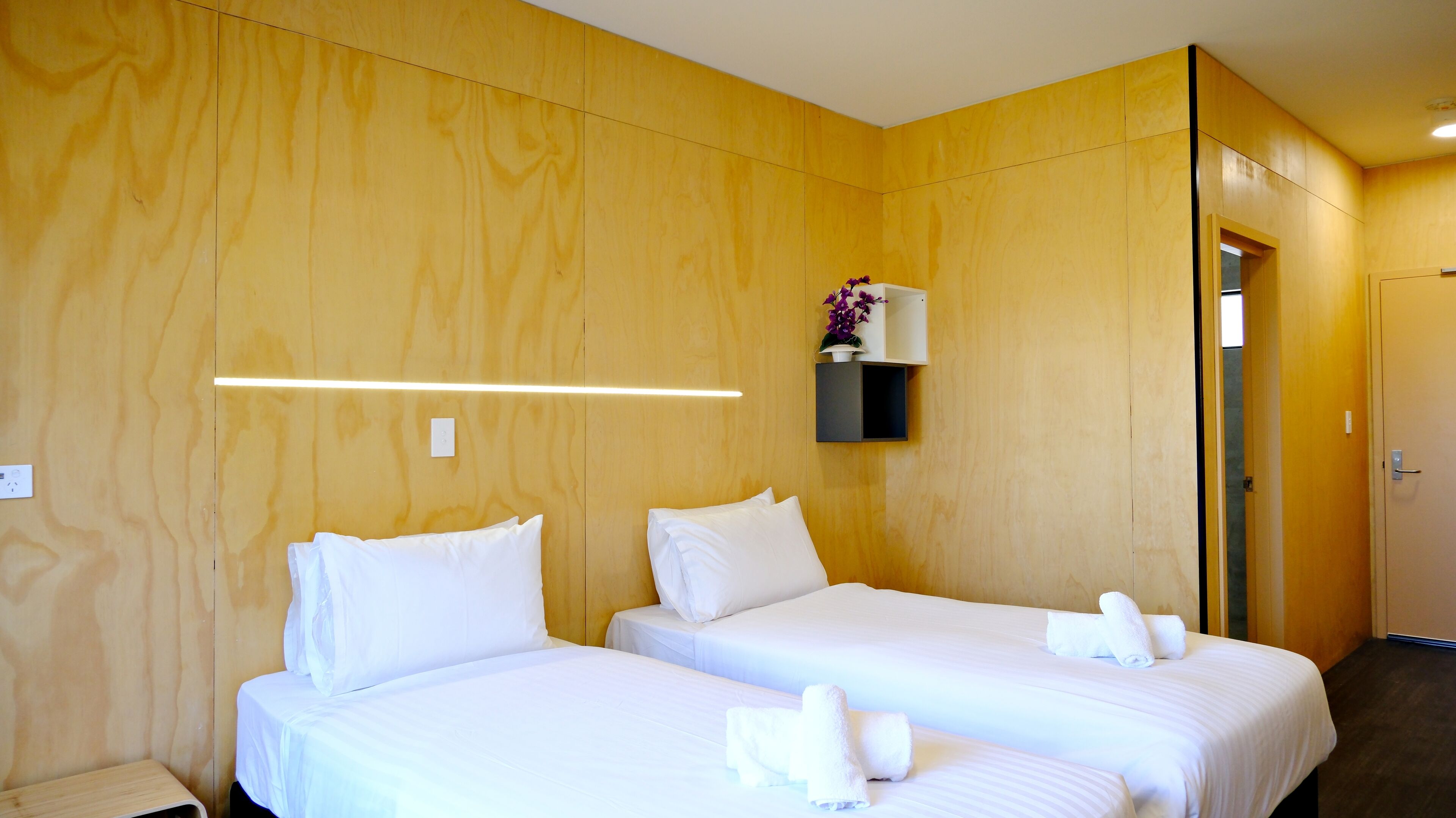 A hotel room with two beds, wooden paneling, a small shelf with a vase, and a door to the right.