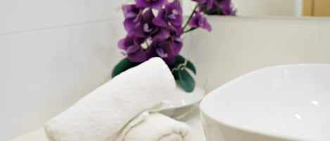 Une salle de bain avec un lavabo blanc, une serviette blanche et des fleurs violettes dans un vase.