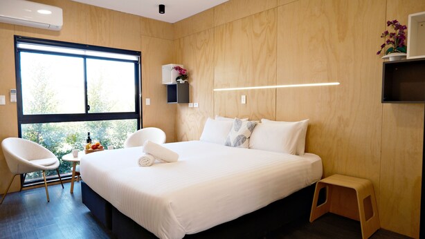 A modern hotel room with a large bed, a wooden headboard, a small desk with a chair, and a window with a view of greenery. - Gallery Hotel Manjimup (Manjimup)
