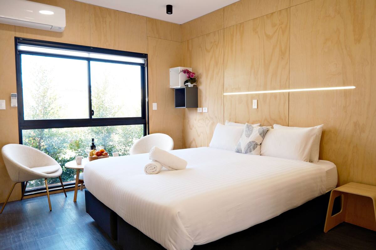 Une chambre d’hôtel moderne avec un grand lit, une tête de lit en bois, un petit bureau avec une chaise et une fenêtre donnant sur un paysage verdoyant.