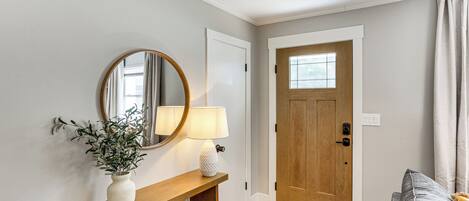 A hallway with a wooden console table, a white vase with a plant, a round mirror, and a lamp. A wooden door with a window is visible in the background.