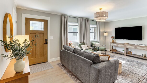 A modern living room with a grey sofa, a wooden door, a large flat-screen TV, and a chandelier. - Modern Retreat w/ Fire Pit Near Harrison Bay! (Chattanooga)