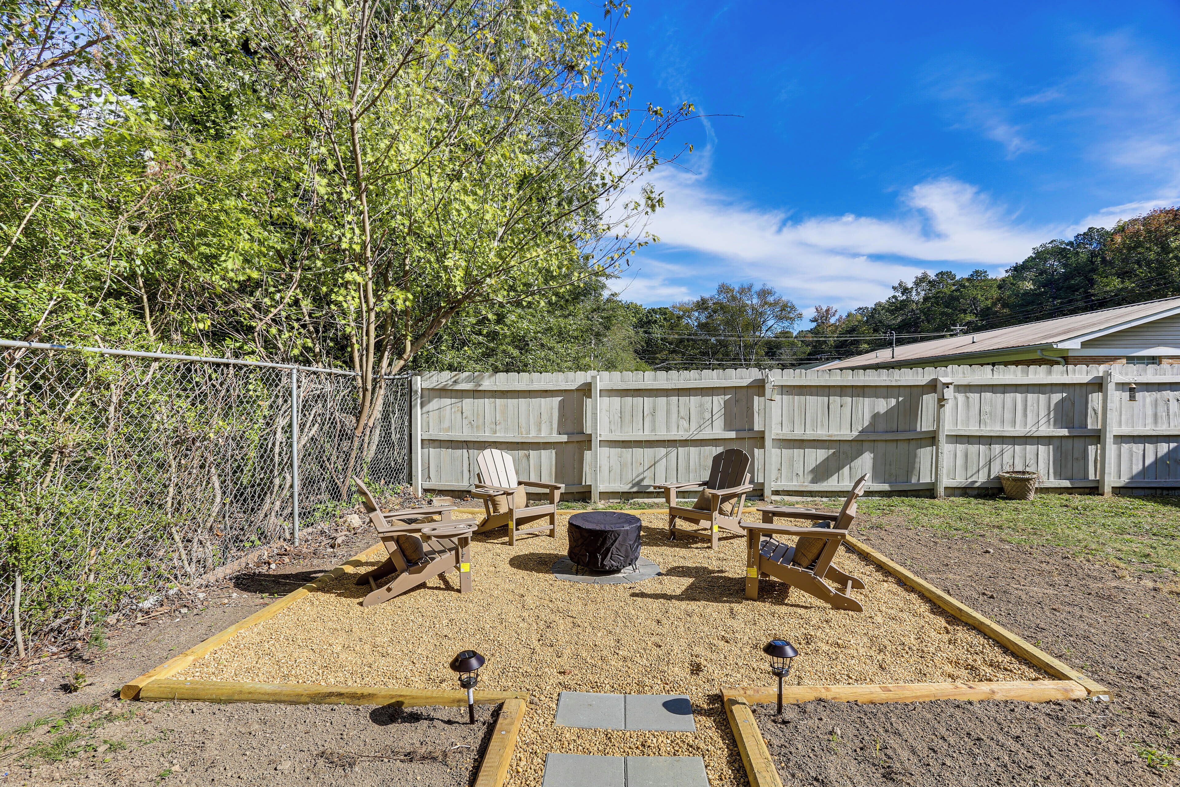 A backyard with a fire pit, wooden chairs, and a fenced area.