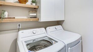 A laundry area with a front-loading washer and dryer, wooden shelves above, and a white cabinet. - Modern Retreat w/ Fire Pit Near Harrison Bay! (Chattanooga)