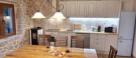 A rustic kitchen with stone walls, wooden beams, white cabinets, and a wooden dining table set for two with a glass of wine and a plate of food.