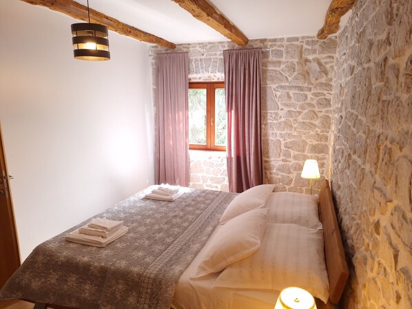 A bedroom with a stone wall, a wooden ceiling beam, a bed with a patterned bedspread, and a window with curtains.