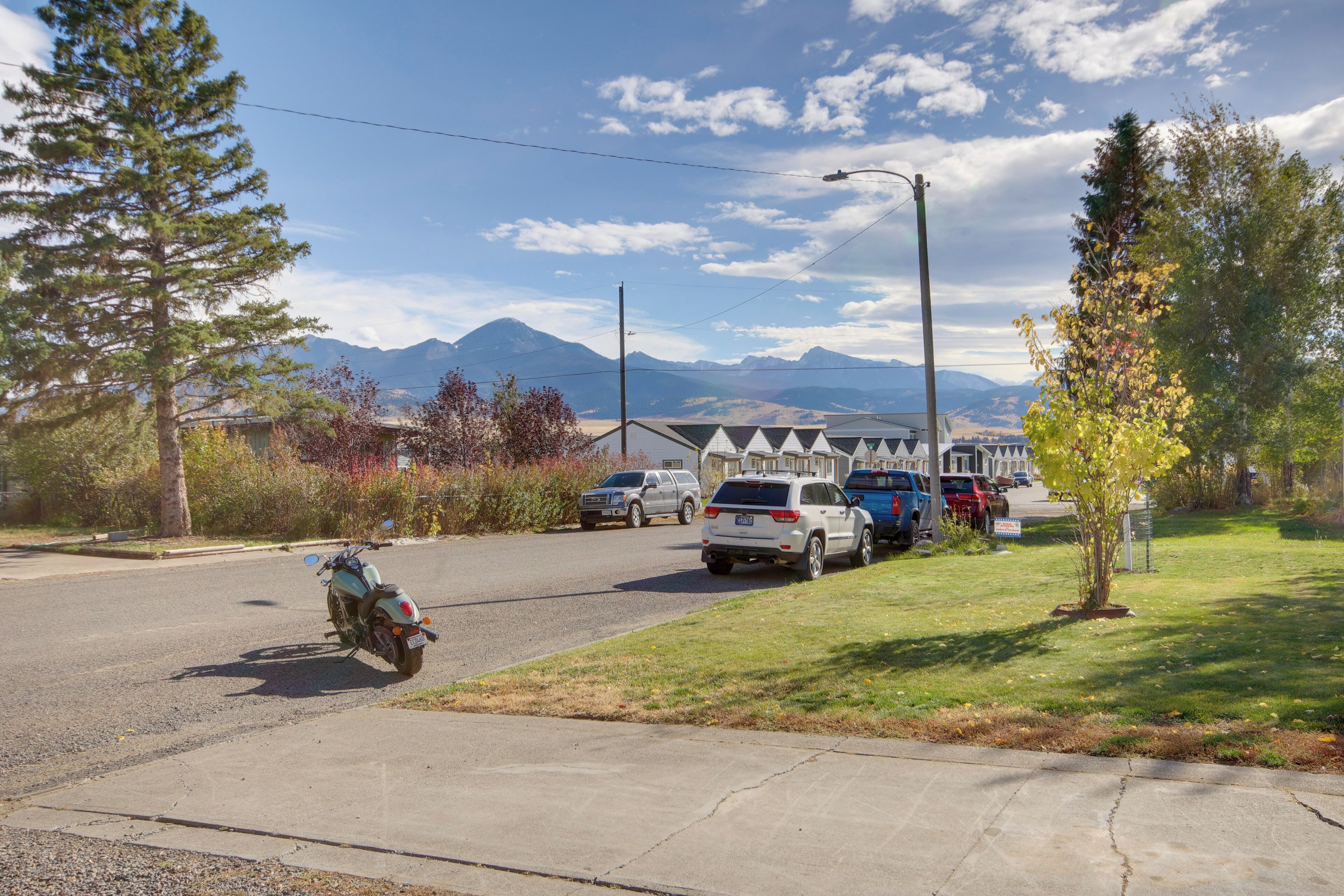 A residential street with parked cars and a motorcycle, houses in the background, and mountains in the distance.