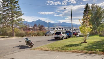 A residential street with parked cars and a motorcycle, houses in the background, and mountains in the distance.