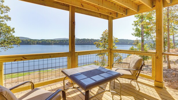 Eine Holzterrasse mit Tisch und Stühlen, die auf einen See mit Hügeln im Hintergrund blickt.