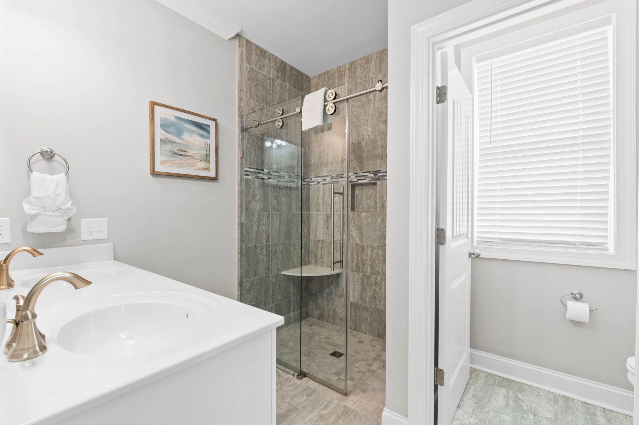 A bathroom with a white sink, gold faucets, a glass shower enclosure, and a framed picture on the wall.