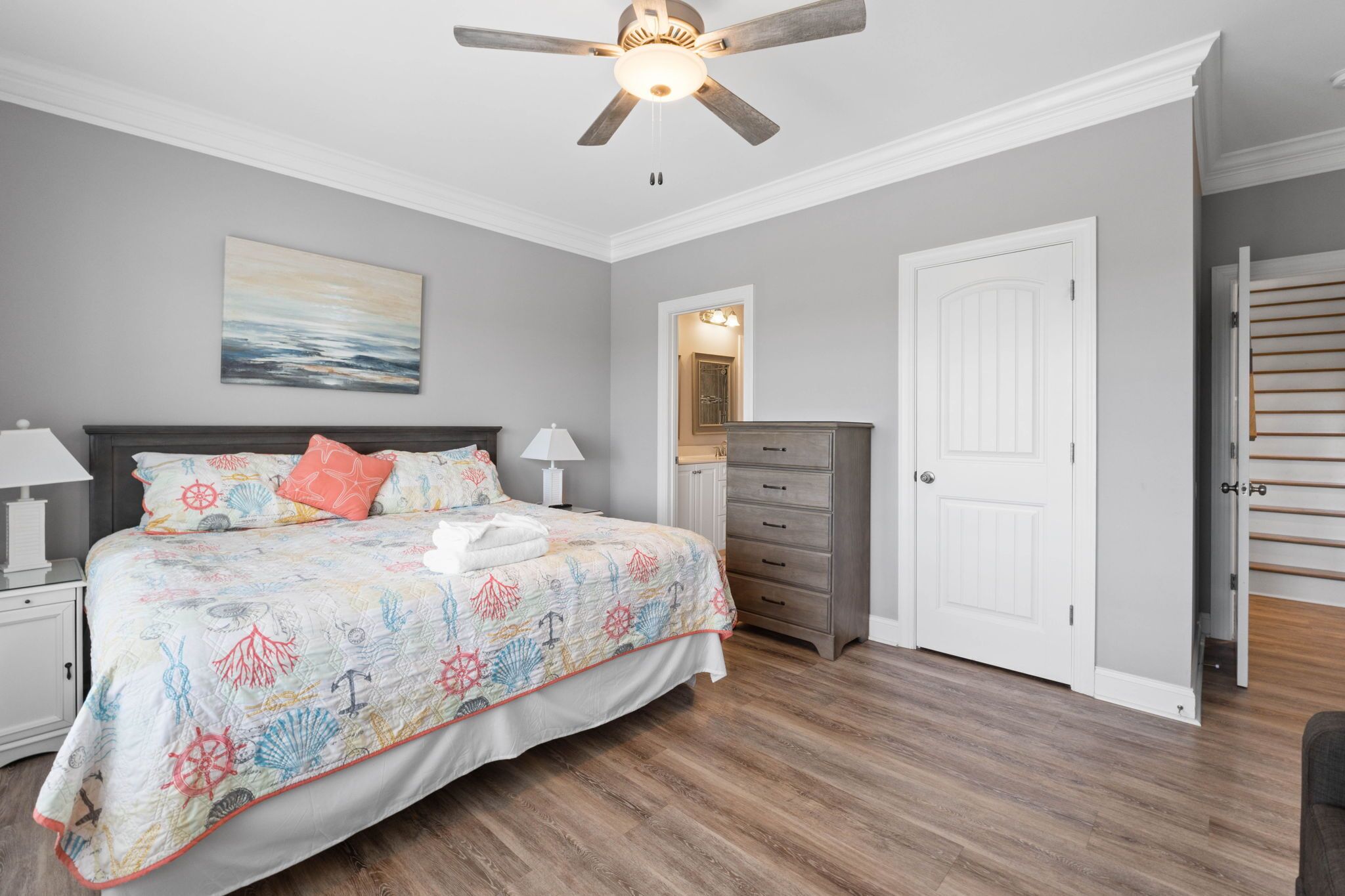 Un dormitorio con cama, mesitas de noche, cómoda, ventilador de techo y una puerta que da a otra habitación.