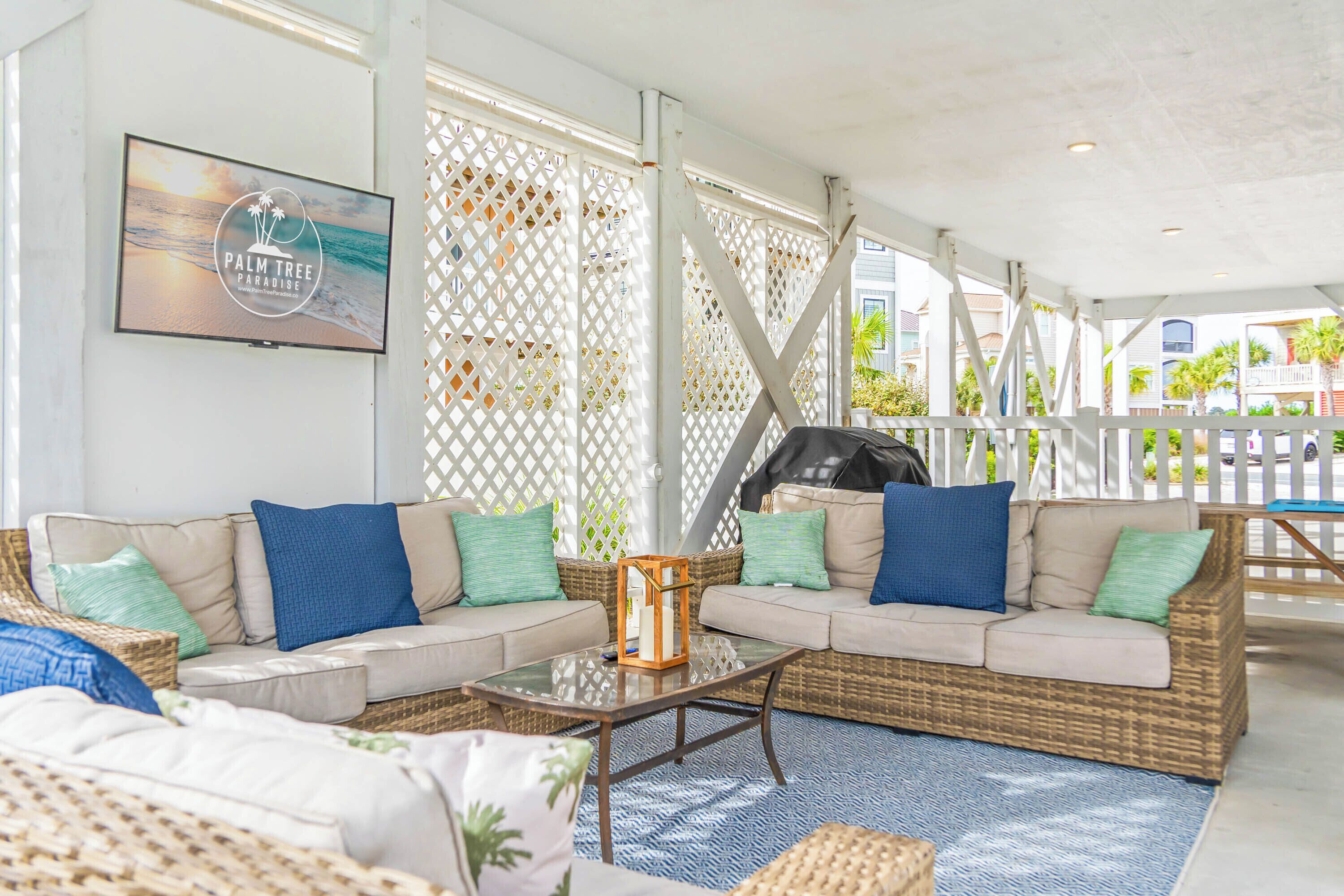 Un área de estar al aire libre techada con mobiliario de mimbre, una mesa de centro de vidrio y una pantalla de televisión que muestra 'PALM TREE PARADISE'.