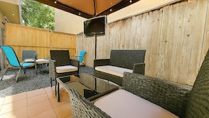 An outdoor patio with wicker furniture, a flat-screen TV mounted on the wall, and a wooden fence in the background.