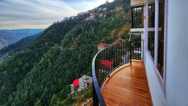 A balcony with a wooden floor and black railing overlooking a mountainous landscape with buildings. - Royal Cedar Shimla (Shimla)