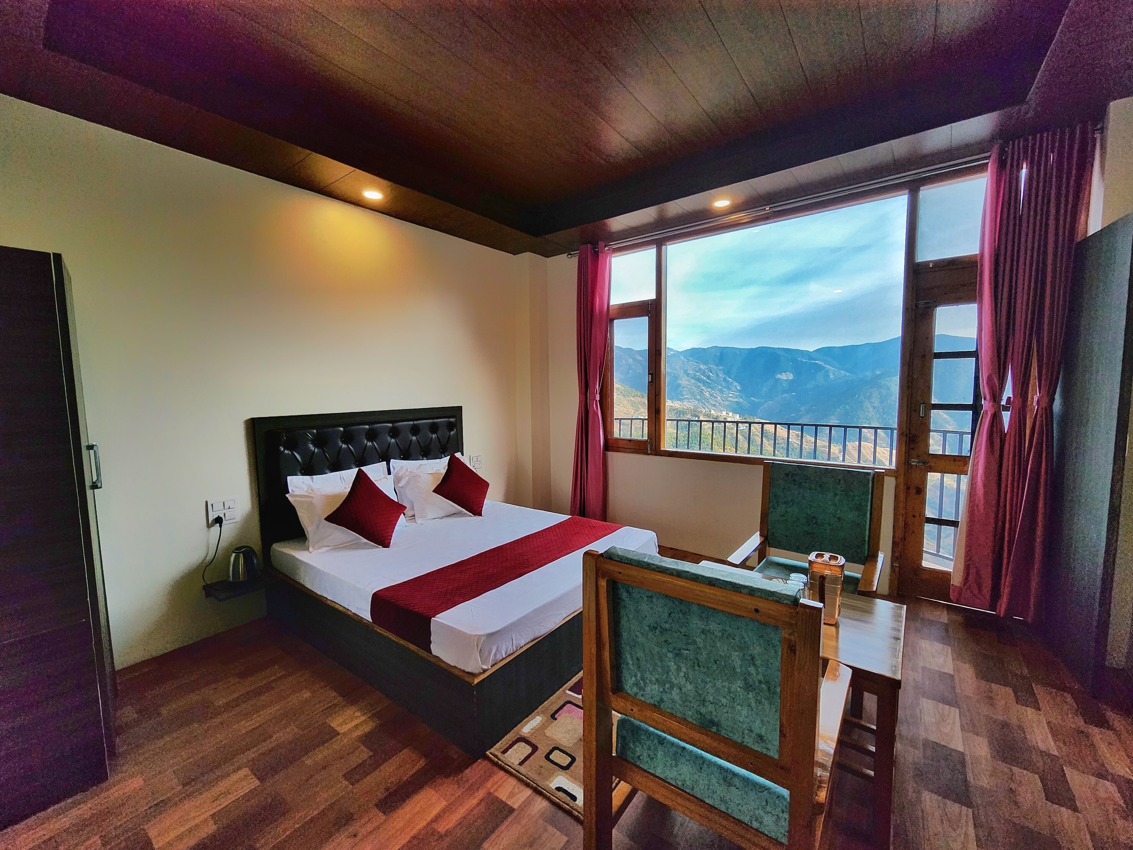 A hotel room with a bed, a desk, a chair, a window with a mountain view, and a wooden floor.