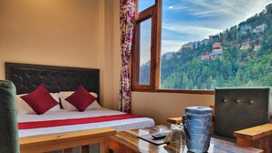 A bedroom with a bed, a window with a view of a forested hillside, a wooden table, and a chair. - Royal Cedar Shimla (Shimla)