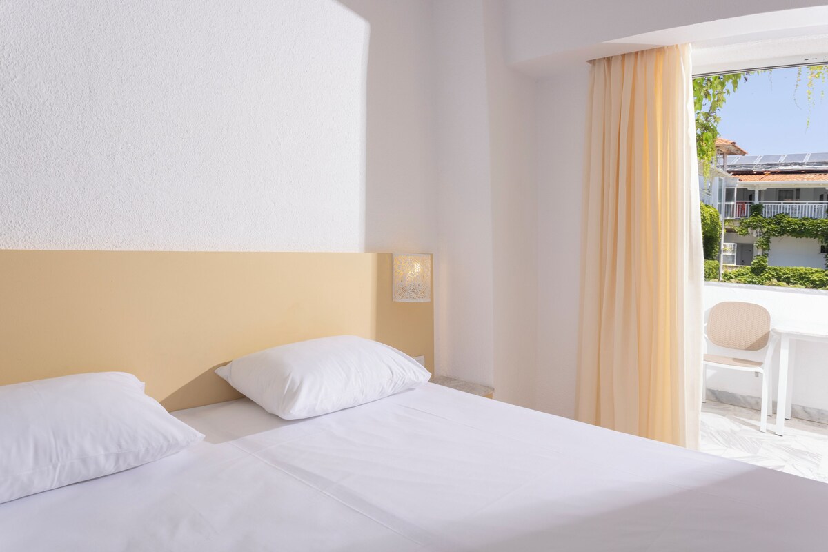 Ein Hotelzimmer mit Bett, Stuhl, einem Fenster mit Vorhängen und Blick auf Gebäude.