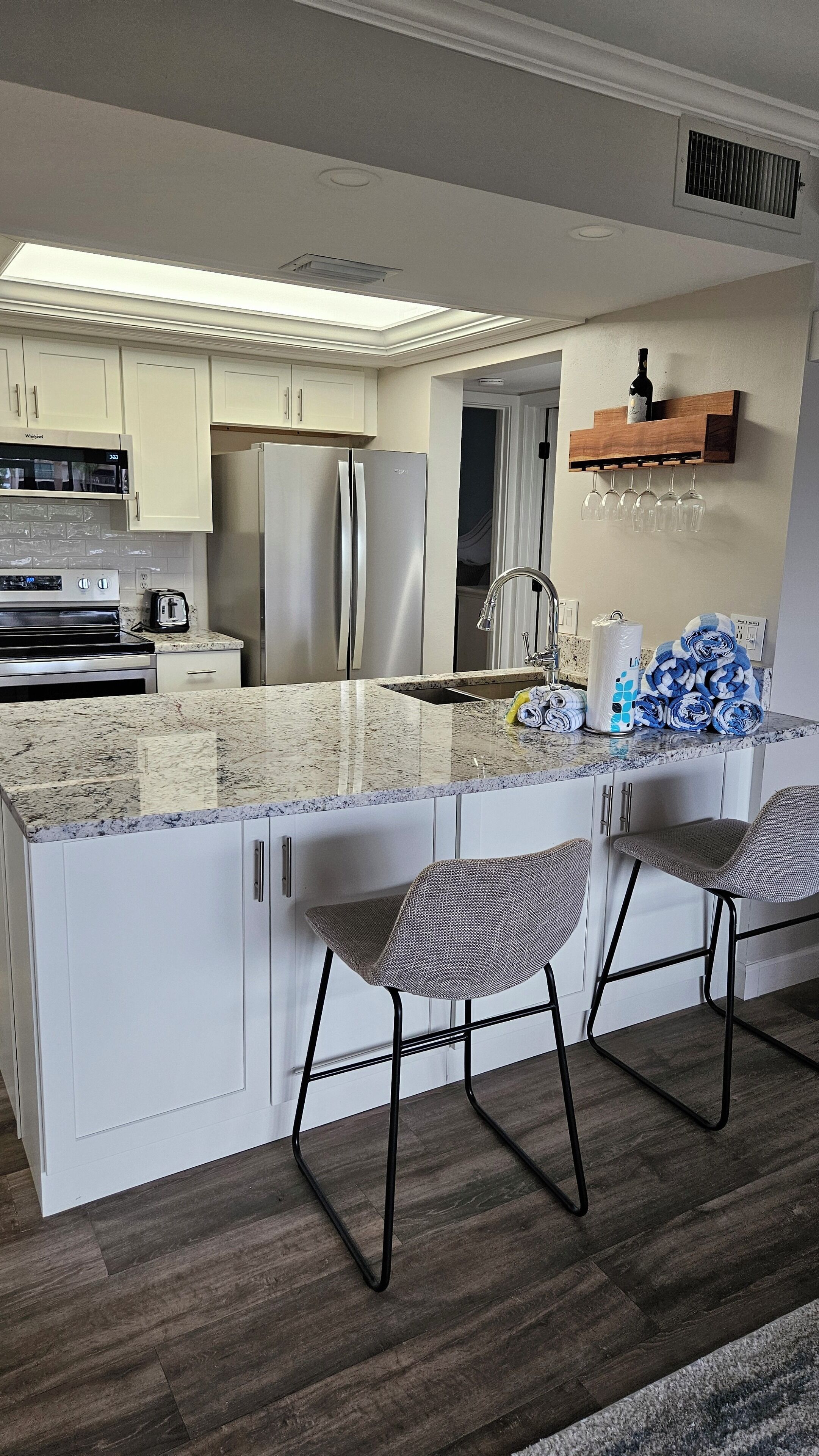 A modern kitchen with white cabinets, stainless steel appliances, a marble countertop, and two bar stools.