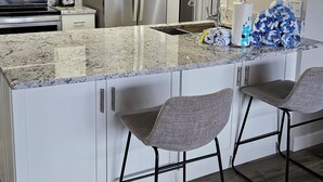 Una cocina moderna con armarios blancos, electrodomésticos de acero inoxidable, encimera de mármol y dos taburetes de bar.