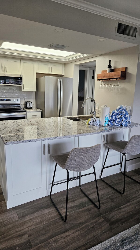 A modern kitchen with white cabinets, stainless steel appliances, a marble countertop, and two bar stools.