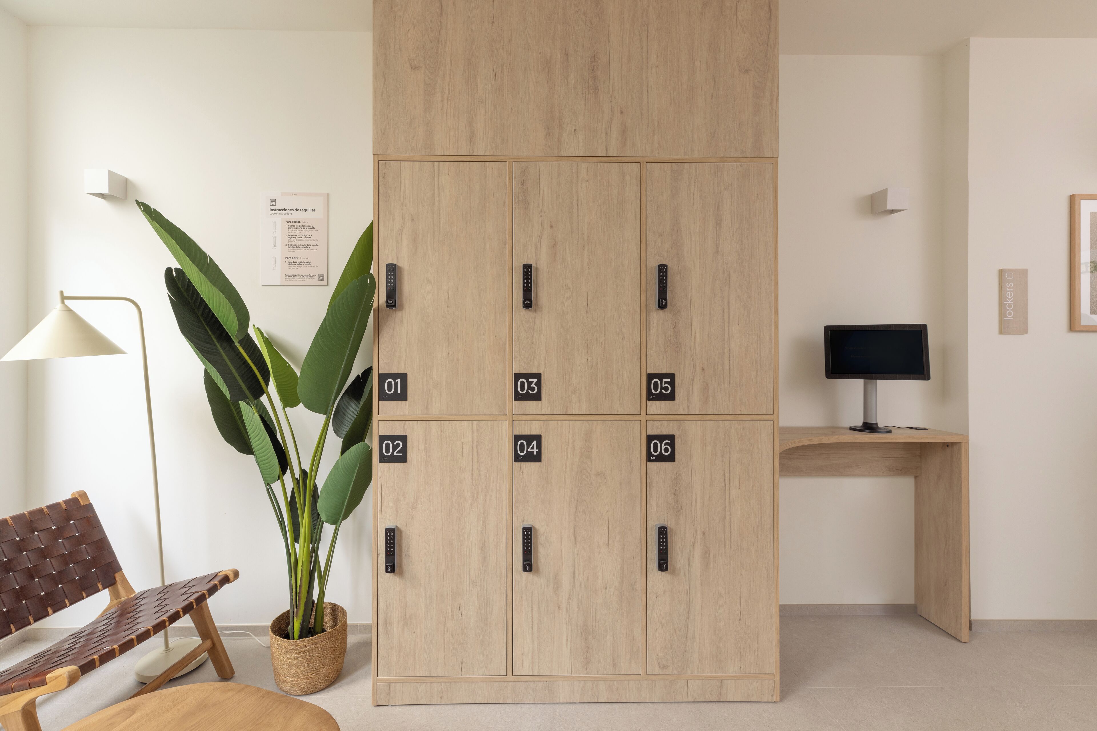 Una habitación con un armario de madera con compartimentos numerados, un escritorio con una computadora, una silla, una lámpara y una planta en maceta.