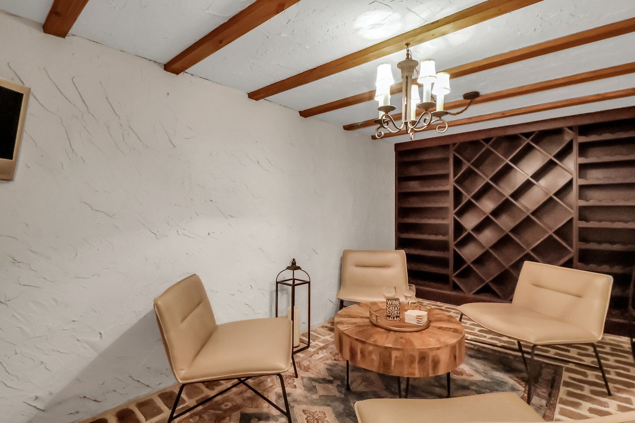 Une pièce avec un mur blanc à la texture intéressante, un plafond en bois et un lustre. On y trouve deux fauteuils beige disposés autour d’une table ronde en bois, sur laquelle se trouve un plateau de collations.