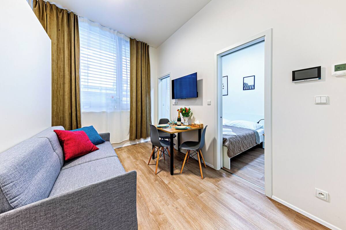 A modern hotel room with a grey sofa, a wooden dining table, a TV, and a bed.