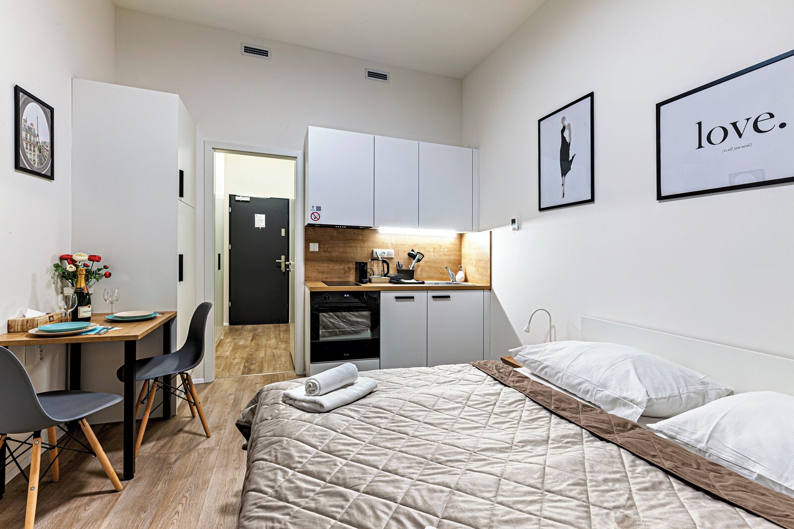 A compact hotel room with a bed, a desk, a kitchenette, and a dining area.