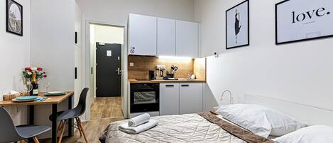 A compact hotel room with a bed, a desk, a kitchenette, and a dining area.