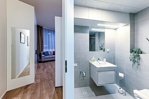 A modern hotel room with a wooden floor, a grey sofa with blue and red cushions, a large mirror, and a white bathroom with grey tiles. - MamaStay Andel apartments (Prague)