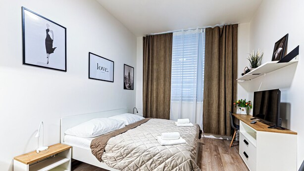 A modern hotel room with a bed, bedside table, TV, and a desk with a chair. - MamaStay Andel apartments (Prague)