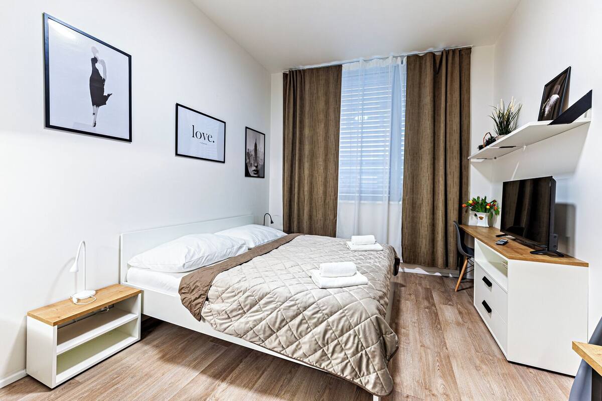 A modern hotel room with a bed, bedside table, TV, and a desk with a chair.
