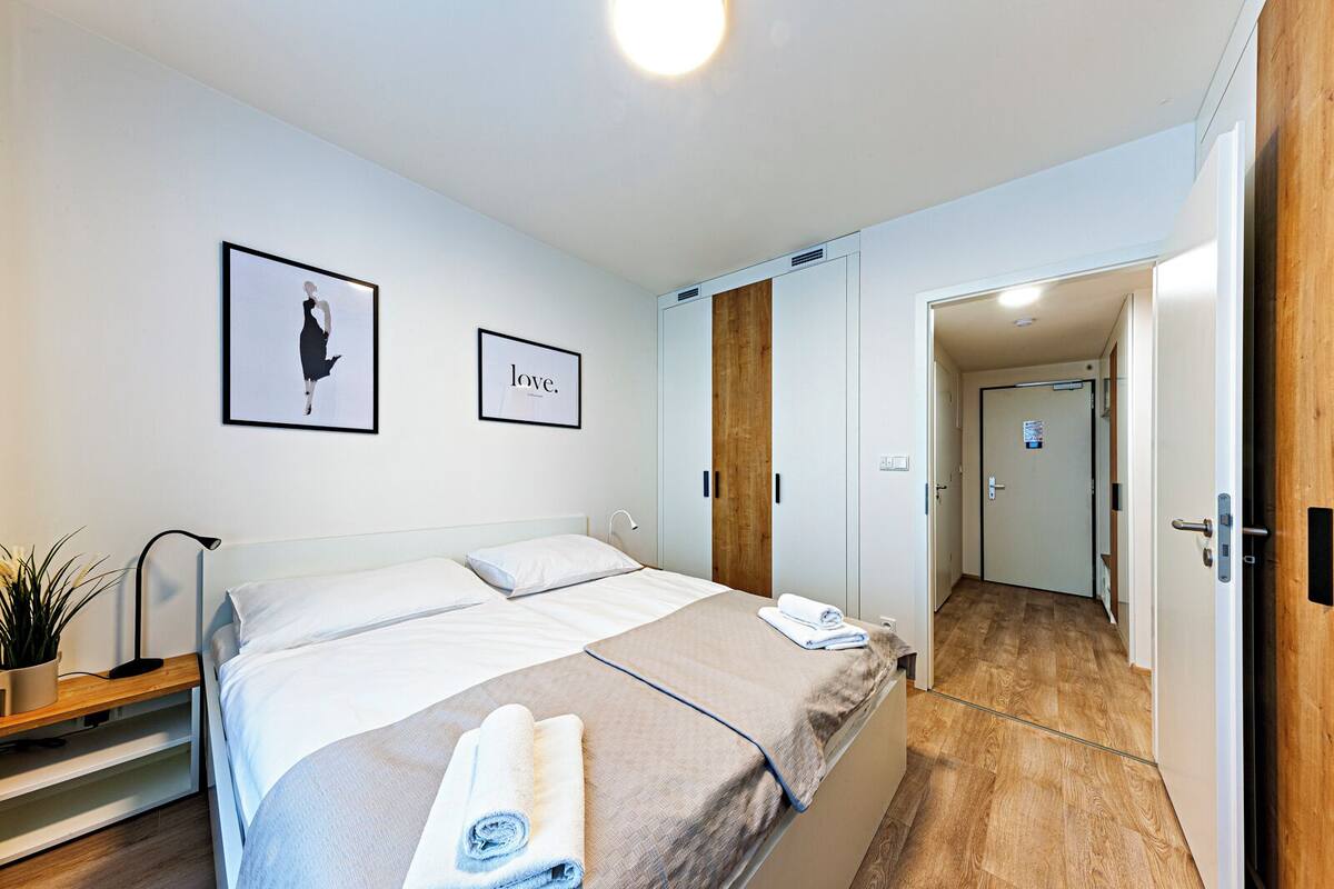 A modern bedroom with a large bed, a bedside table with a lamp, a potted plant, and two framed pictures on the wall.