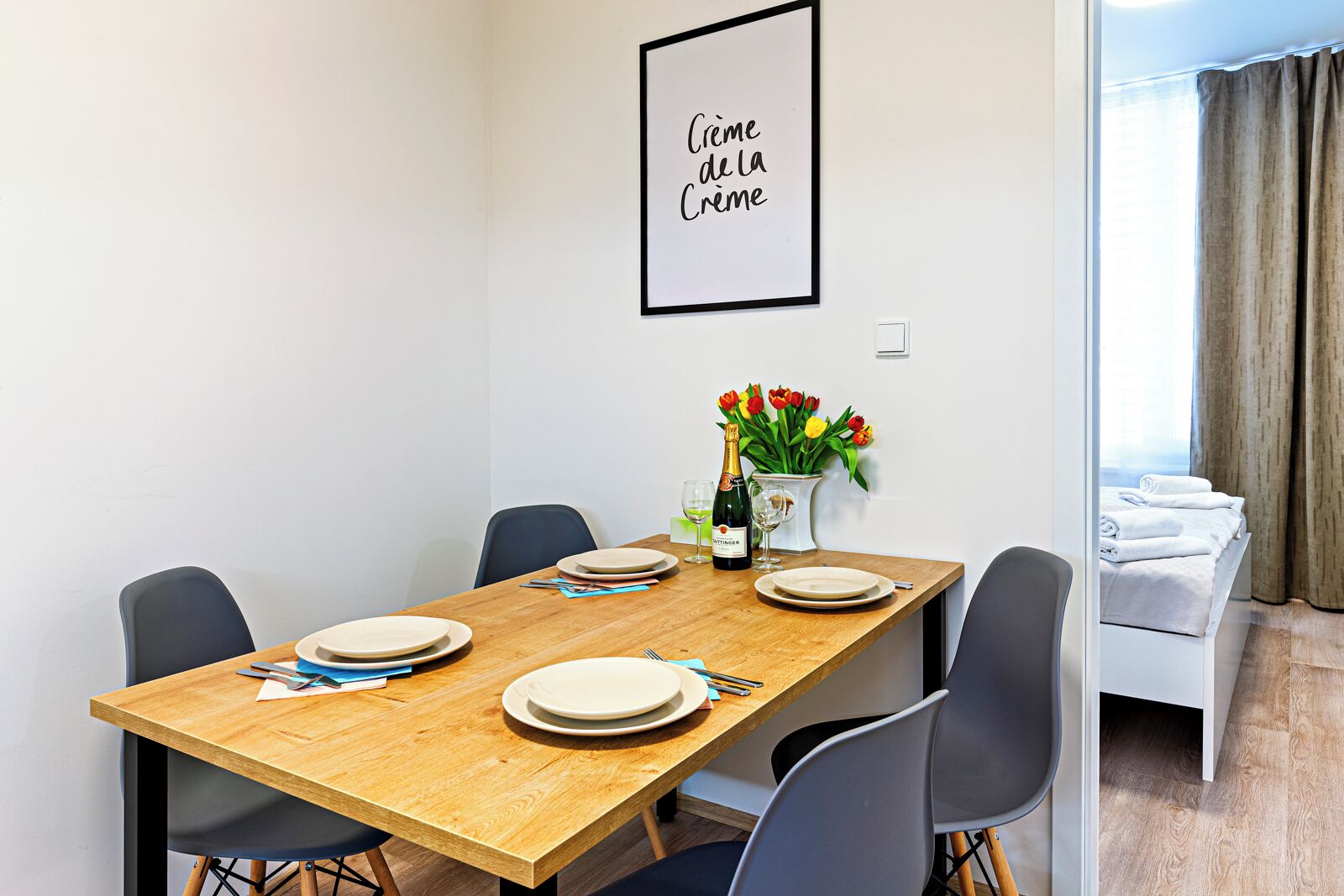 A dining area with a wooden table set for four, a vase of tulips, and a bottle of champagne.