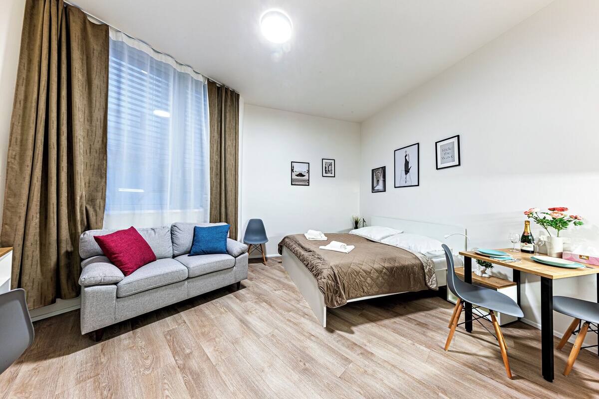 A modern hotel room with a bed, a sofa, a desk with a chair, and a window with curtains.