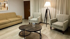 A hotel room with a sofa, two armchairs, a small round table with water bottles, a floor lamp, and a painting on the wall.